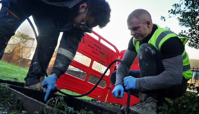 Two technicians performing drainage maintenance, using tools to clear a drain, with a red service vehicle in the background, highlighting emergency drainage solutions for schools.