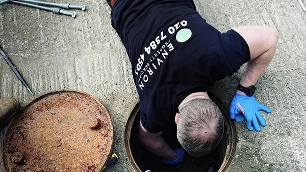 Drainage engineer inspecting a blocked drain, wearing gloves and a branded Environ shirt, with tools and a rusty drain cover nearby.