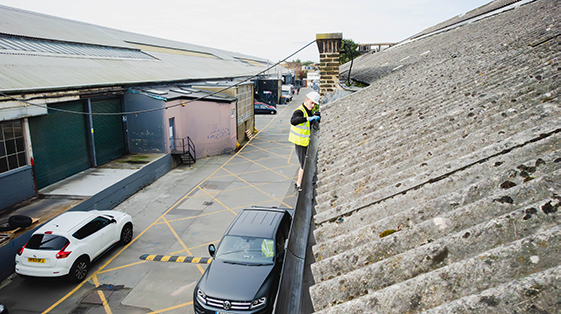 Worker in safety gear cleaning commercial gutter on a building roof, with parked vehicles and industrial setting in the background, emphasizing professional gutter maintenance services.