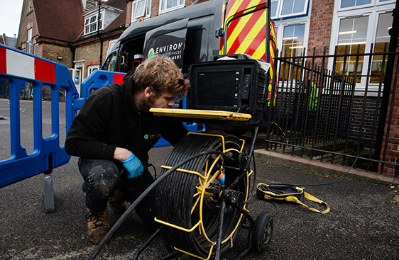 Drain unblocking technician using equipment for school drainage services, with Environ Drainage vehicle and safety barriers in background.