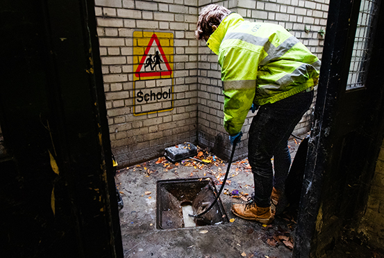 Worker in high-visibility jacket clearing a drainage system in a school environment, with a caution sign indicating "School" on the wall.