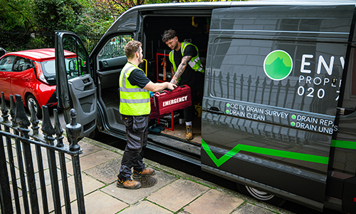 Two drainage engineers in high-visibility vests loading emergency equipment from a van labeled "Environ Drainage Services," parked near a residential area, emphasizing urgent drainage solutions in London.