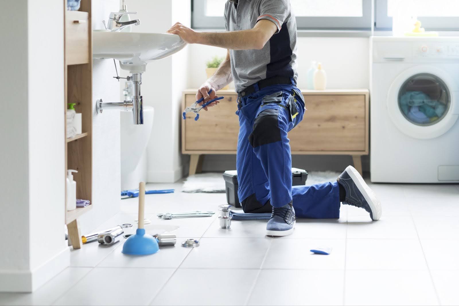 Plumber kneeling and repairing a sink with tools in a modern bathroom, illustrating emergency drainage services.